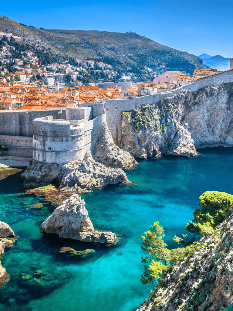 Ancient city walls overlooking a turquoise sea with rocky cliffs and colorful rooftops in a coastal town.