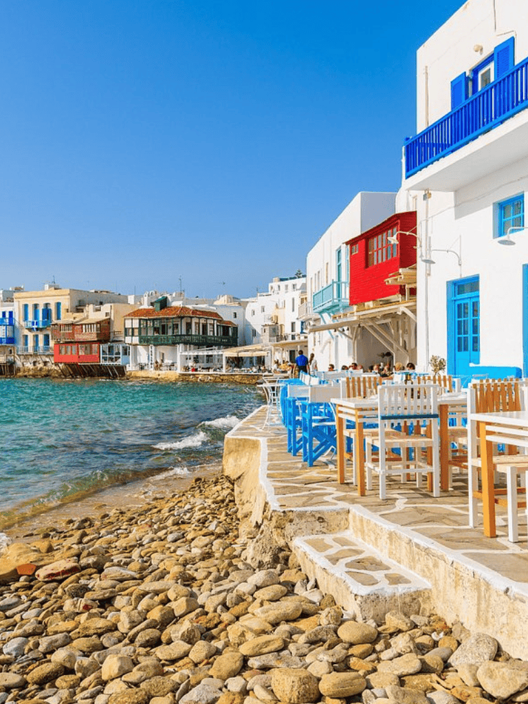 Colorful seaside buildings with outdoor dining overlooking a rocky shoreline in a popular Greek island.
