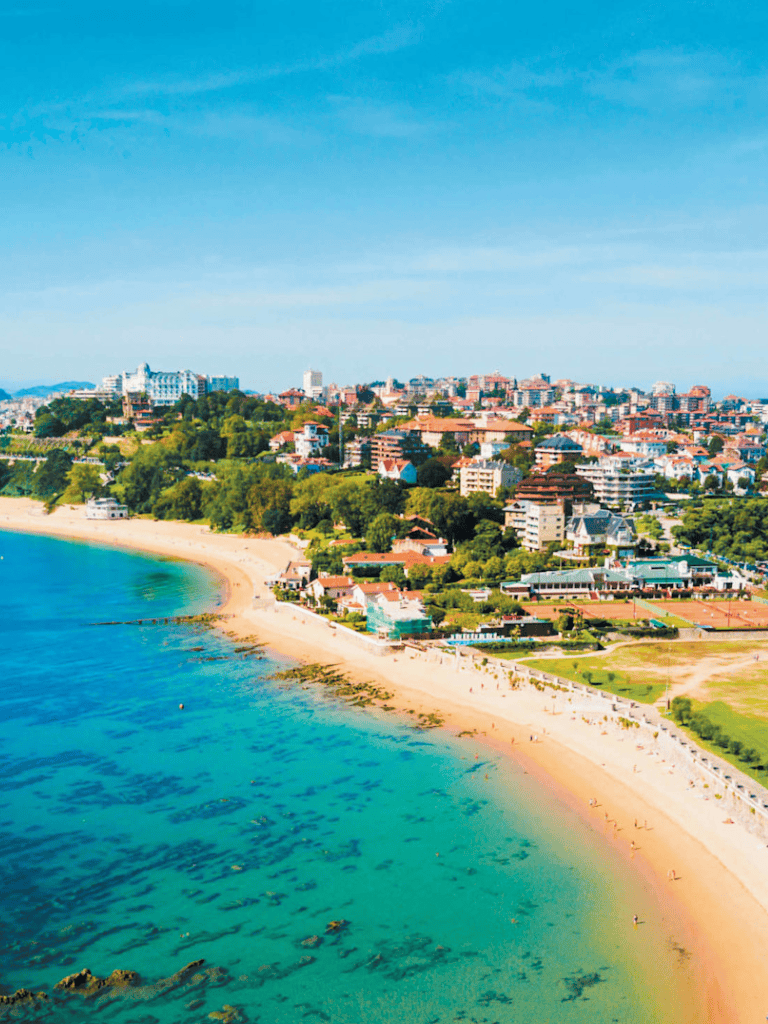 Aerial view of a coastal city with beaches, buildings, and lush greenery under a clear blue sky.