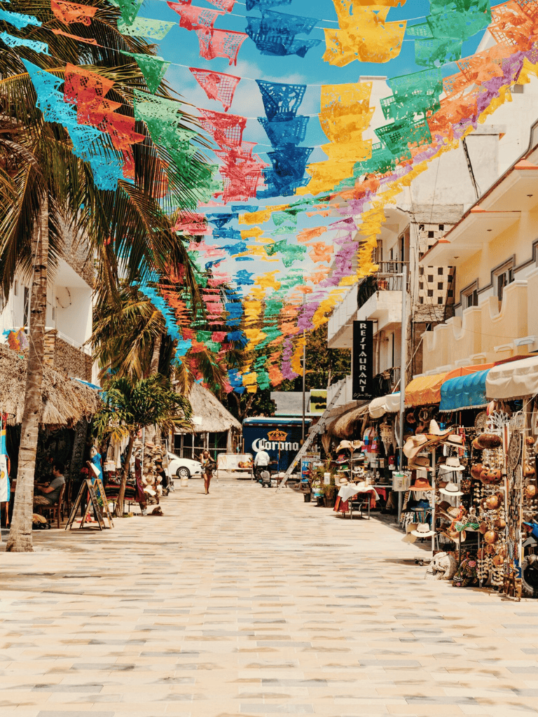 Colorful Mexican decorations hang above a lively market street with shops and local vendors.