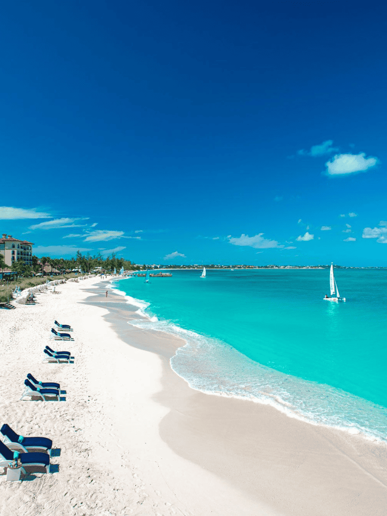 Pristine white sandy beach with turquoise waters and sailboats under a bright blue sky.