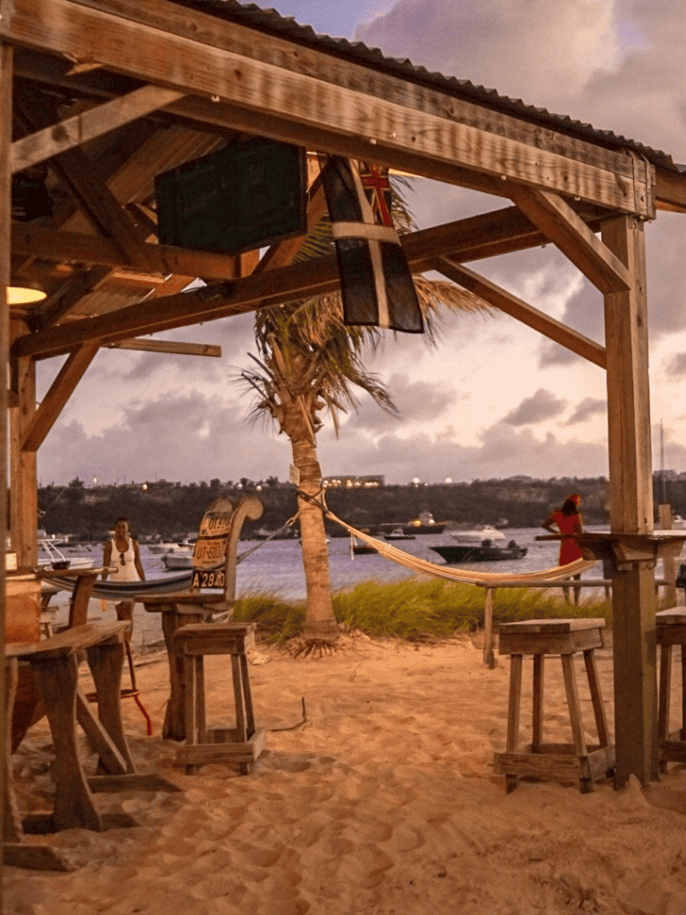 Relaxed beachside tiki bar with boats in the background, tropical palm, and evening sky.