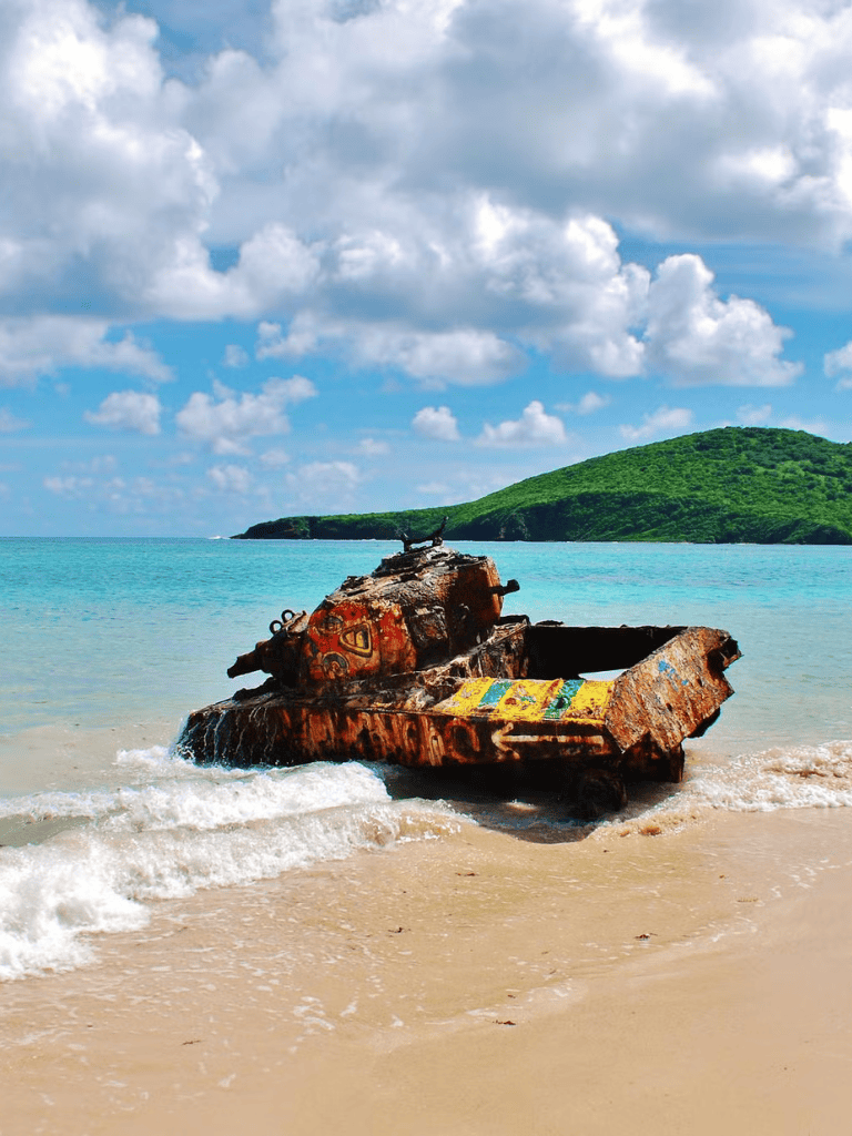 Rusty tank wreck on tropical beach with green hill and blue sky in background, abandoned vehicle photography.