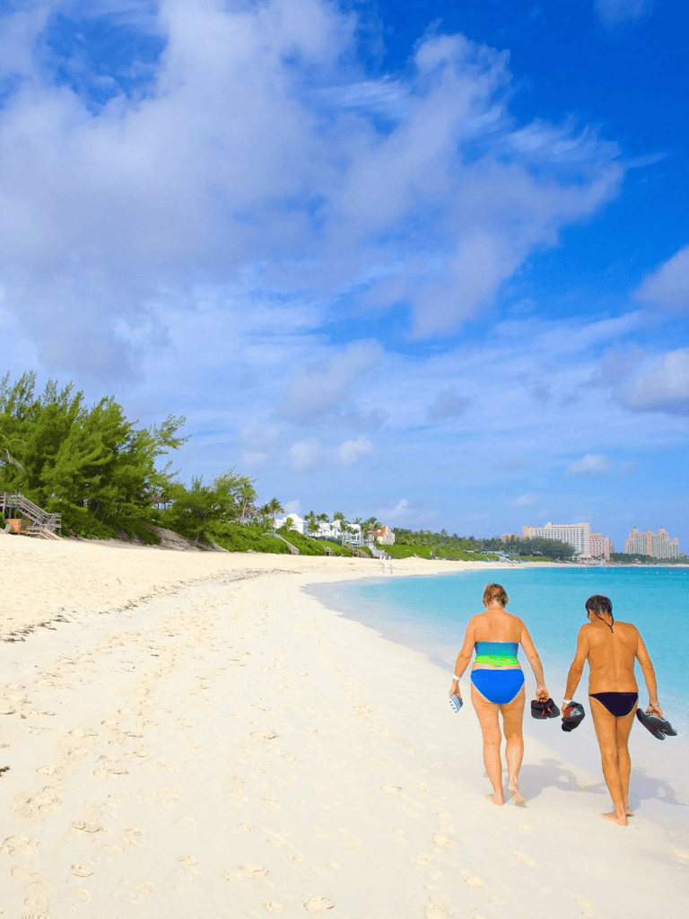 Relaxing beach scene with two women walking along sandy shoreline under blue sky, sunny weather, and lush greenery.