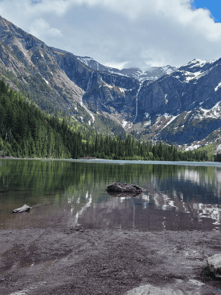 Serene mountain lake with lush pine forest and snow-capped peaks in the background.
