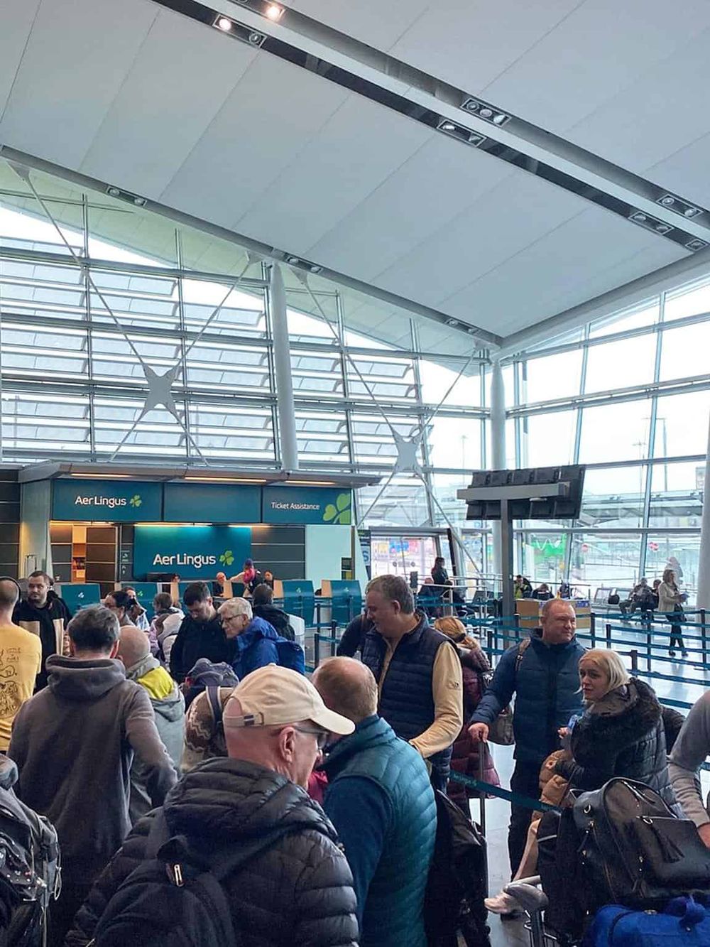 Crowded airport terminal with travelers at check-in counter, modern architecture, and large windows.