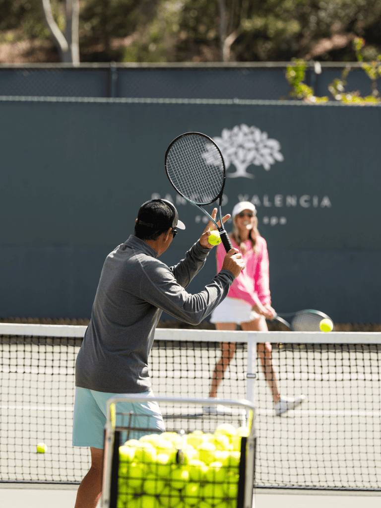 Tennis players practicing on an outdoor court with a basket of tennis balls nearby.