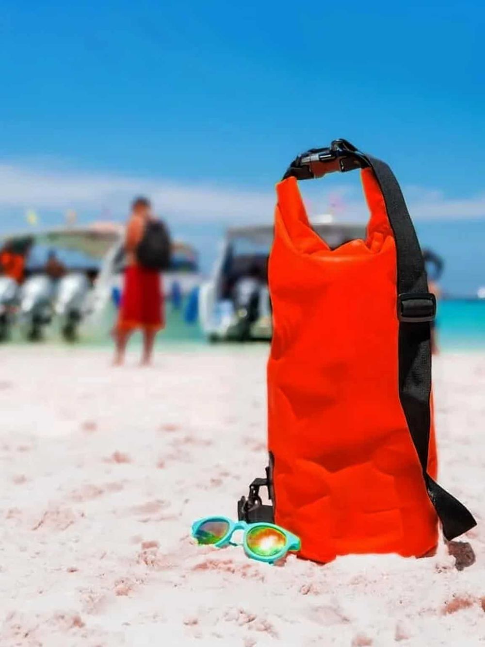 Dry bag for beach water activities and sunglasses on sandy shore with boats and people in the background.