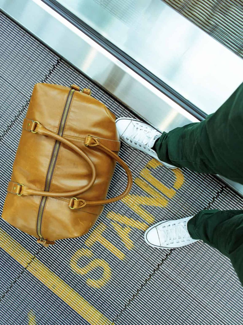 Travel planning at airports, traveler with leather bag, white sneakers, green pants on moving walkway.