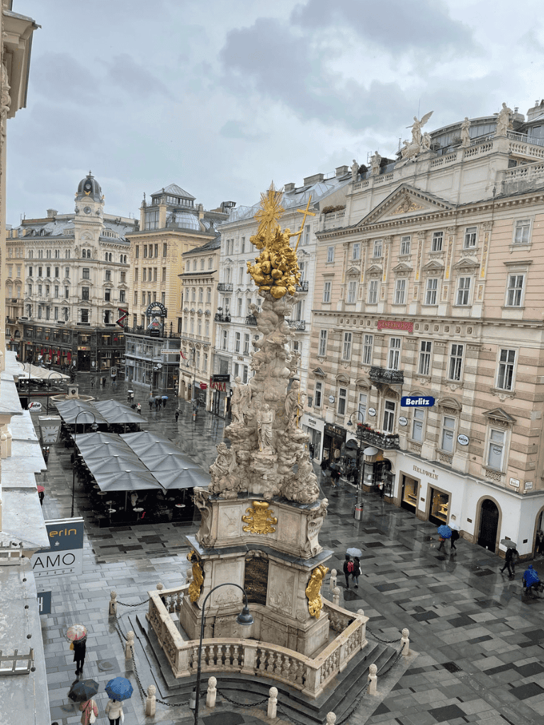 Intricate baroque street fountain in Vienna, Austria, with decorative sculptures and gold accents, under cloudy skies.