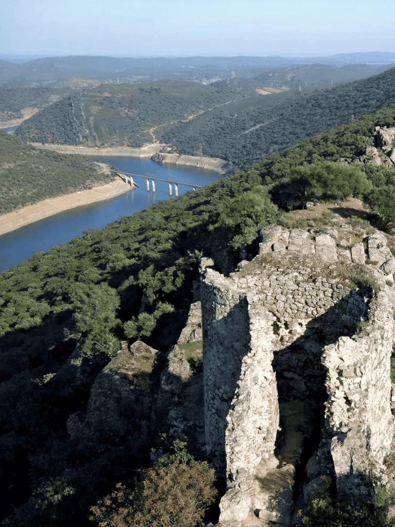 Vast canyon view with river and bridge, lush green surrounds, ancient stone ruins in foreground.