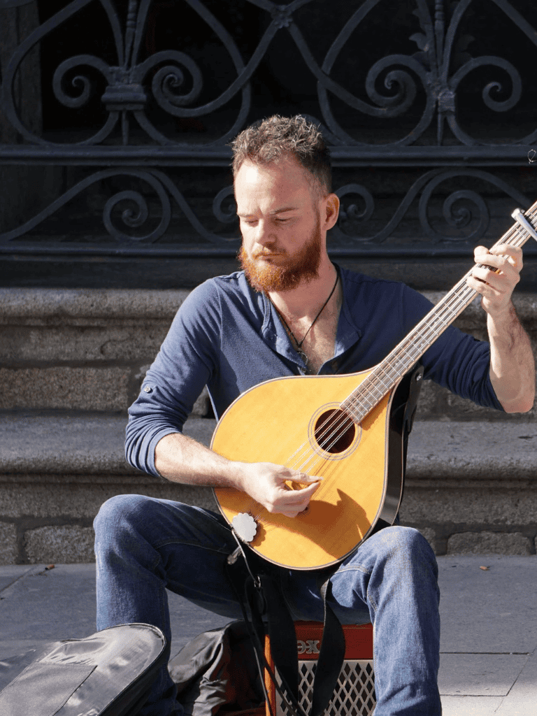 Musician playing acoustic guitar outdoors on city street with stairs and wrought iron fence in background.