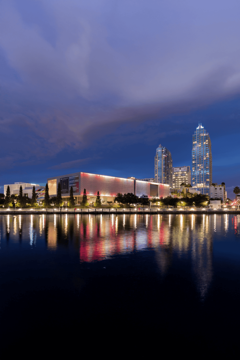 Modern city skyline with tall buildings and vibrant lights reflecting on water.