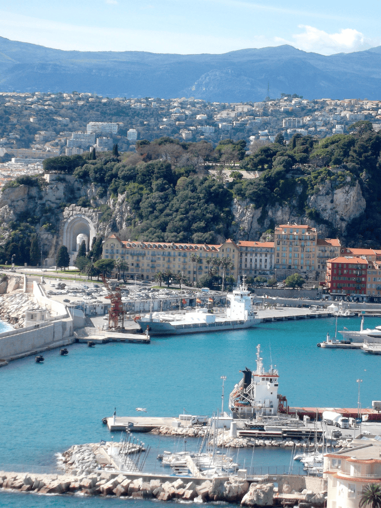 Vibrant harbor scene with yachts and coastal cityscape, hills and mountains in the background.
