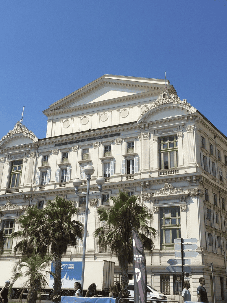 Grand historic building with ornate architecture and palm trees in front, situated in an urban cityscape.