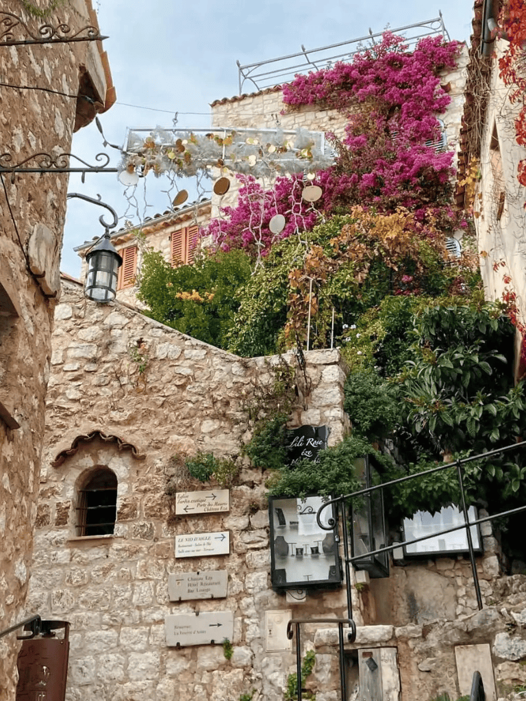 Charming stone alleyway with vibrant bougainvillaea and directional signs in a picturesque village.
