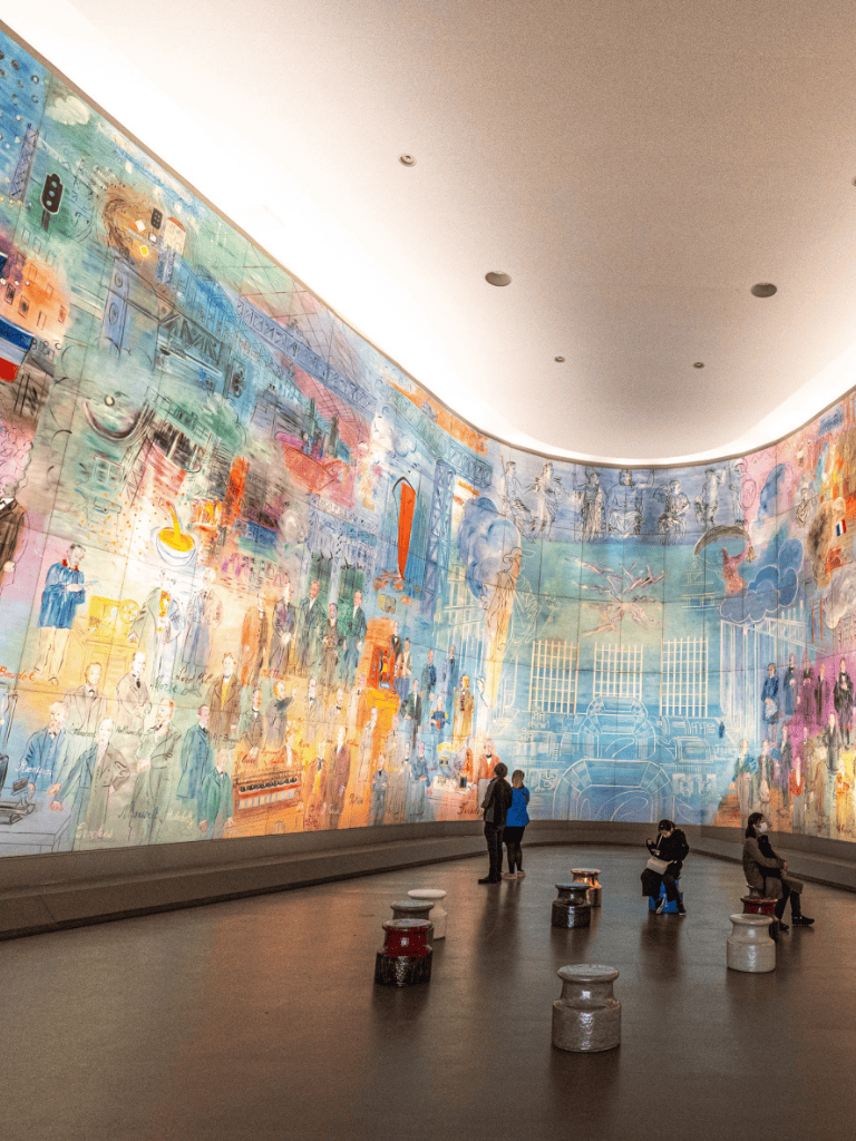 Vivid indoor mural art depicting cityscapes and people, with visitors observing in modern gallery space.