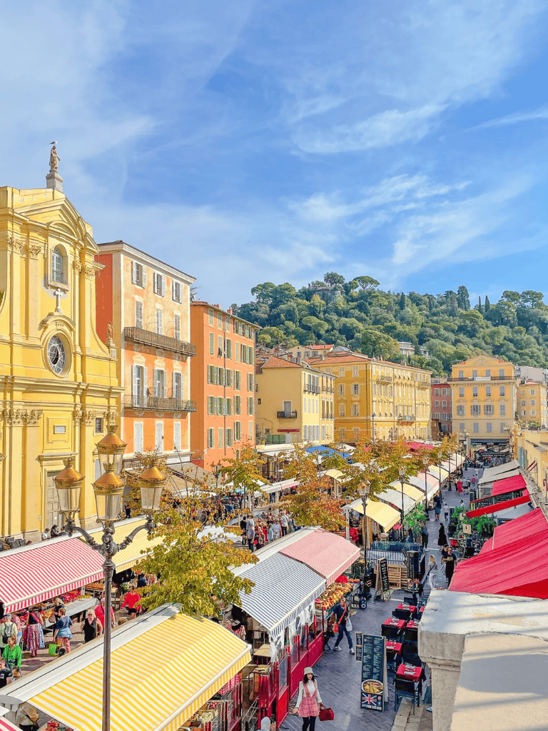 Vibrant outdoor market in a historic European city with colorful buildings and scenic hillside background.