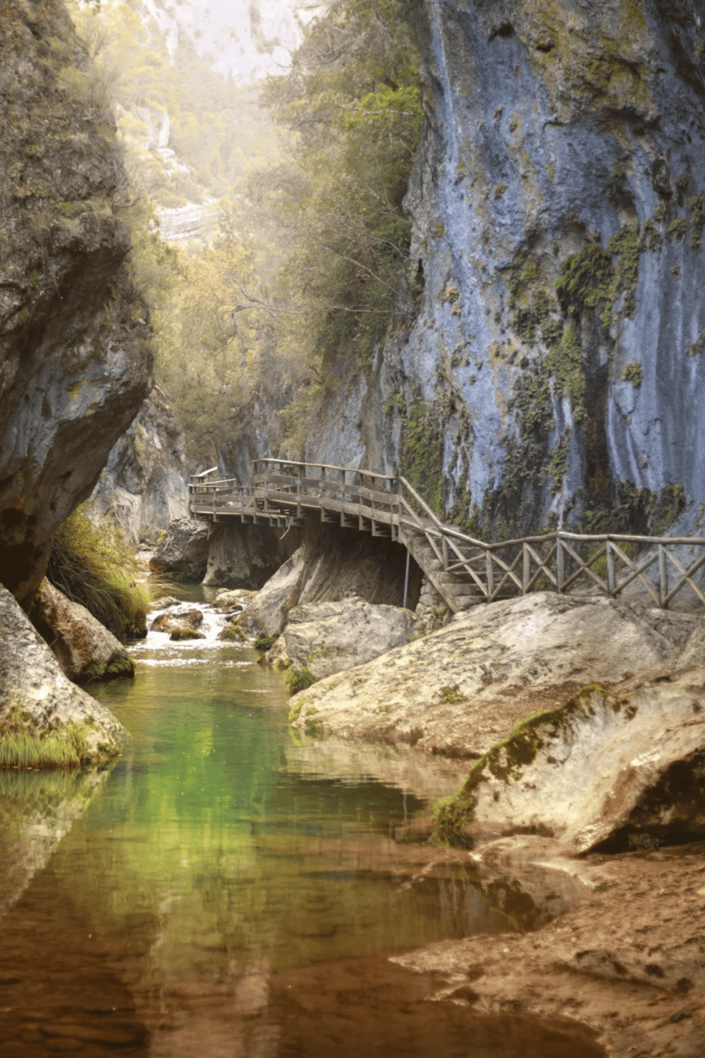 Serene mountain stream with wooden walkway, lush greenery, and rugged cliffs, perfect for nature exploration.