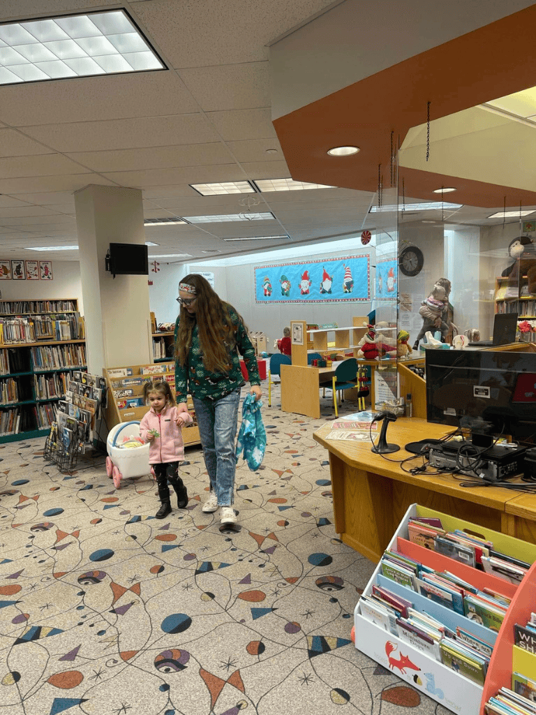 Mom and daughter in a children's section of a library decorated for Christmas with festive displays and holiday books.
