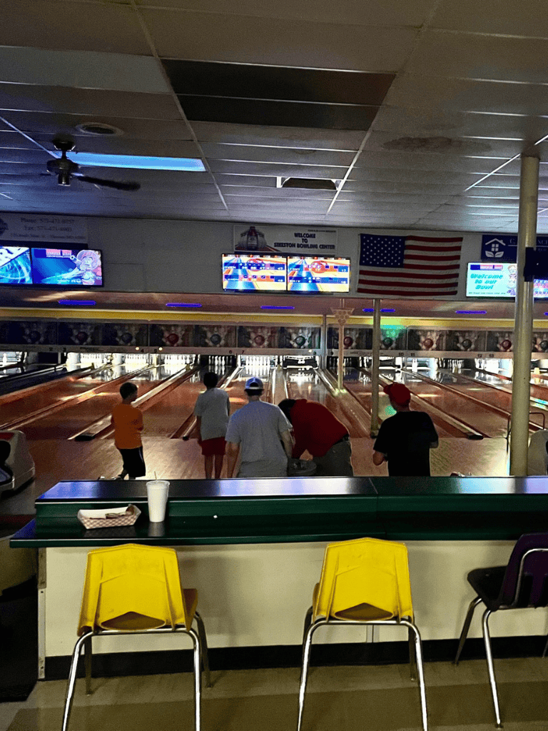 Bowling alley with players and American flag, indoor recreational center with digital scoreboards.