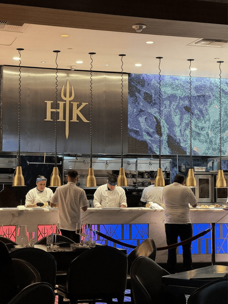 High-end Greek restaurant interior with chefs preparing food at a sleek marble counter, modern lighting, and a decorative wall display.