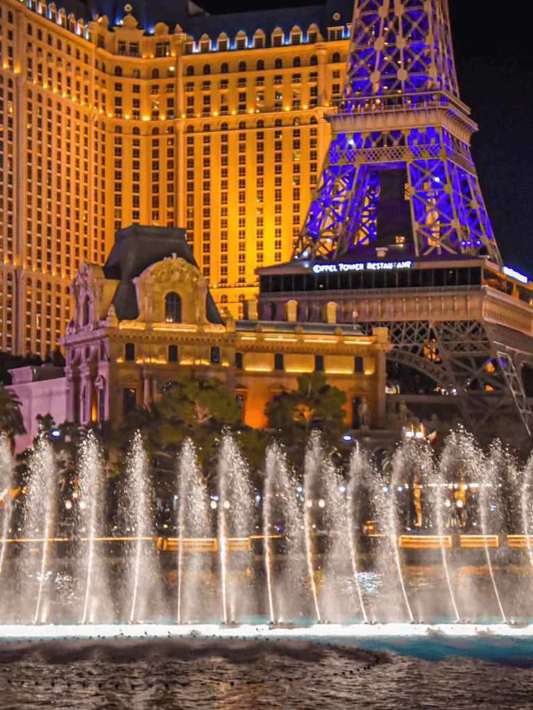 Eiffel Tower replica and Bellagio fountains in Las Vegas at night, illuminated with vibrant lights.