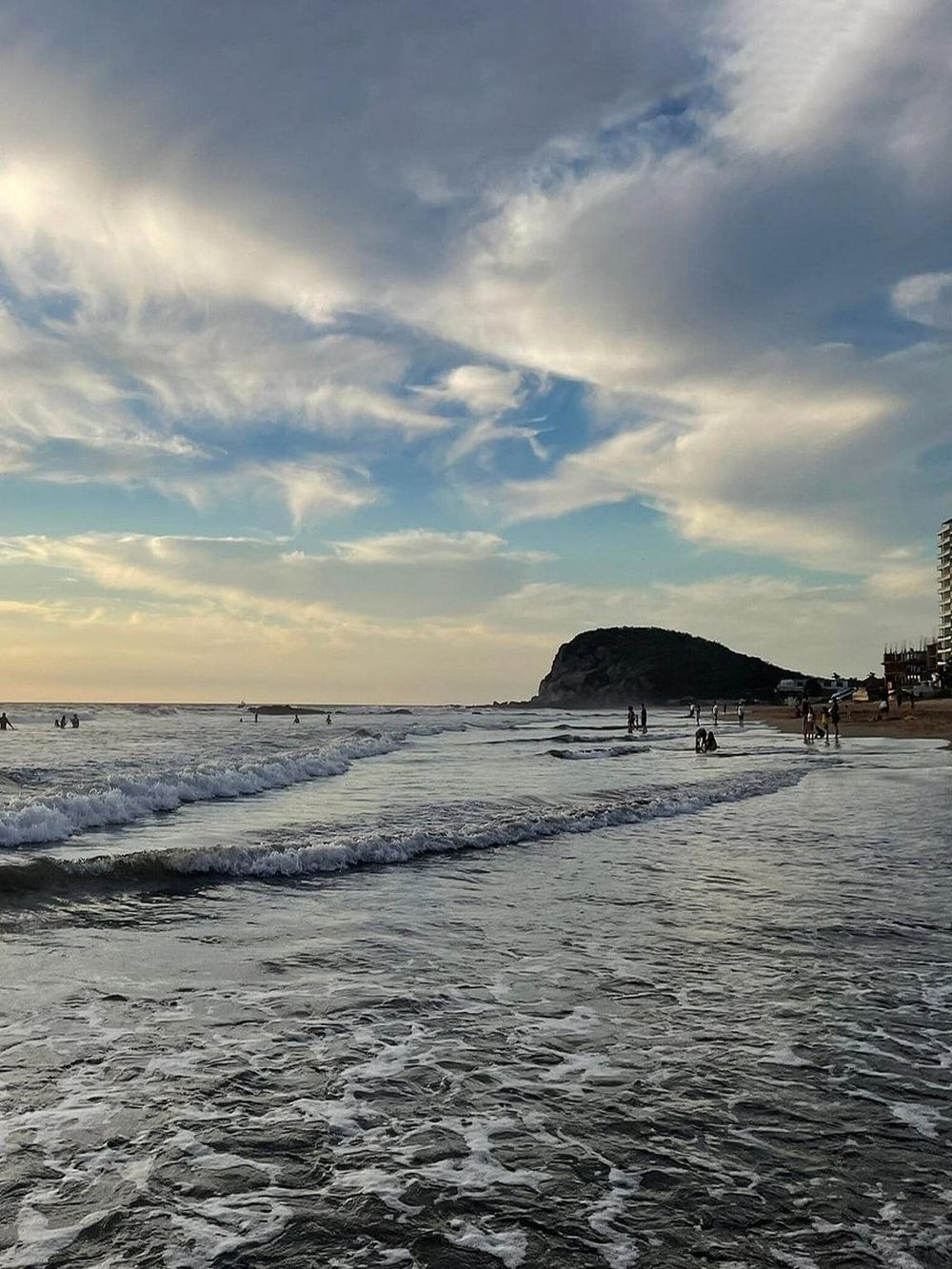 Vibrant beach scene with people enjoying waves and scenic coastal view during sunset.