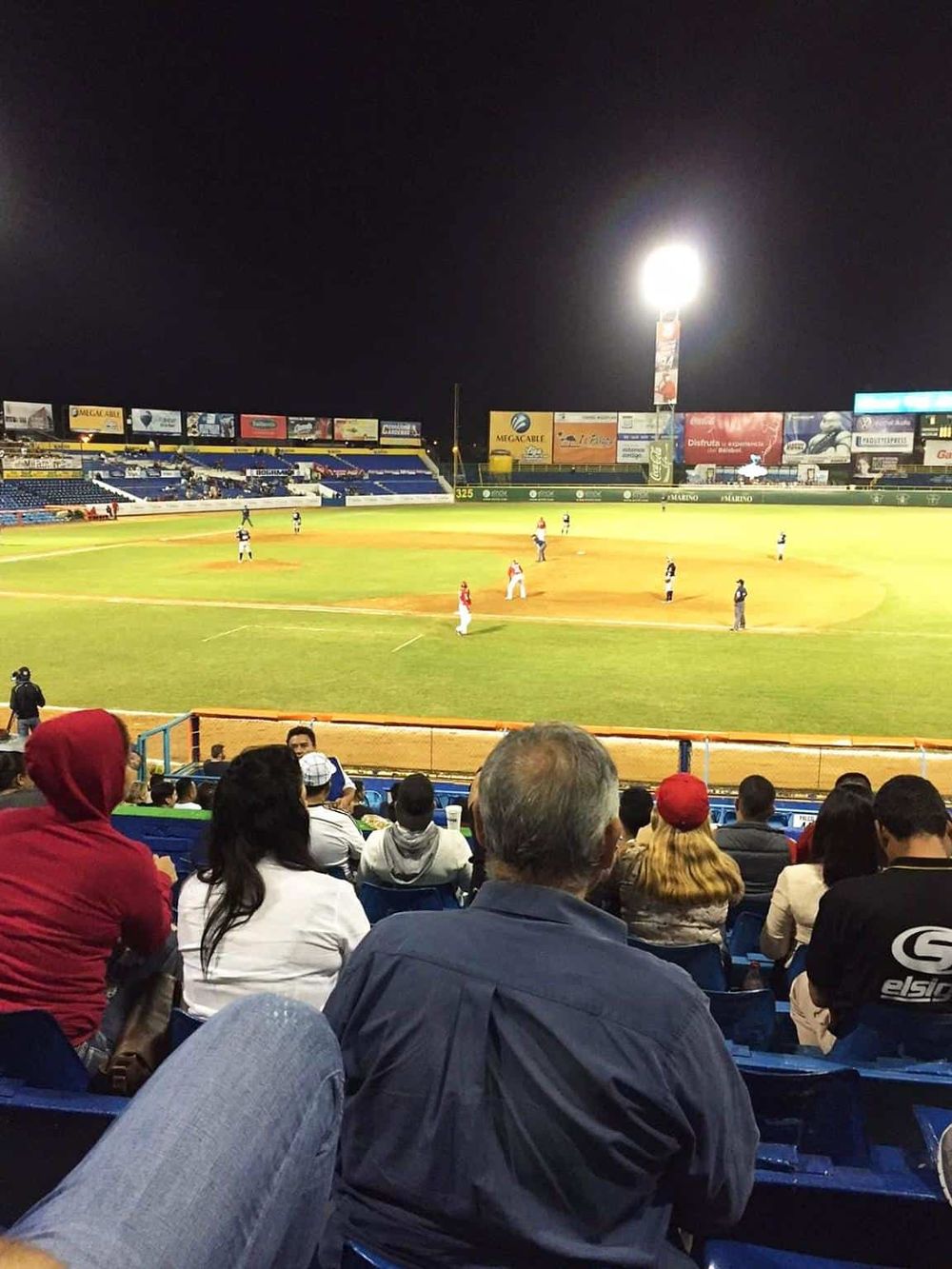 Nighttime baseball game at stadium with fans watching from seats, bright lights illuminating the field, and advertisements on board.