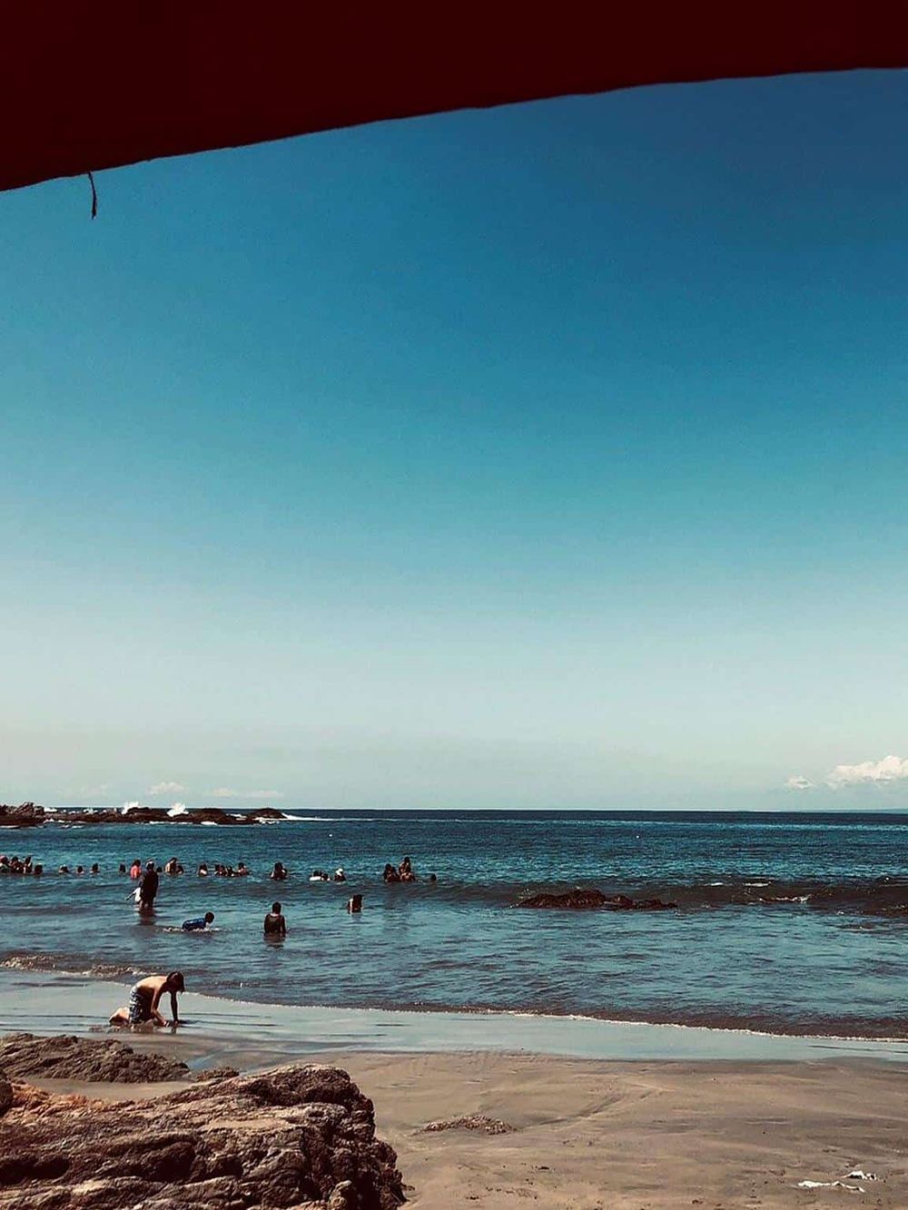 Children playing on beach with ocean waves, sunny sky, and rocky shoreline.