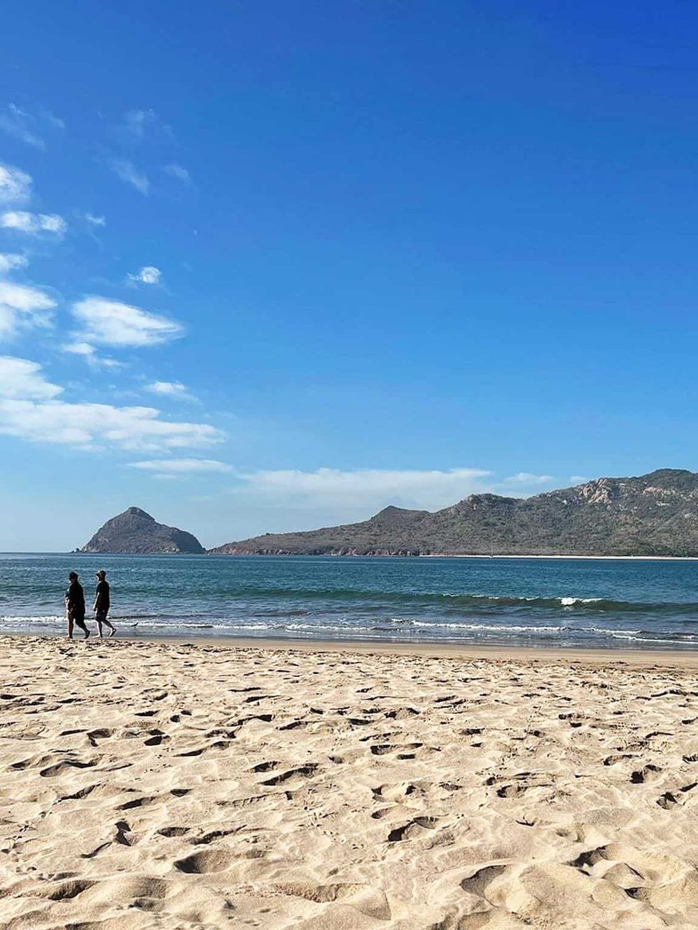 Two people walking on sandy beach with ocean and mountains in background, clear blue sky, scenic coastal landscape.