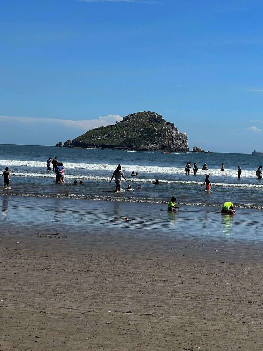 Aerial view of a busy beach with people swimming and playing, with a scenic island in the background.