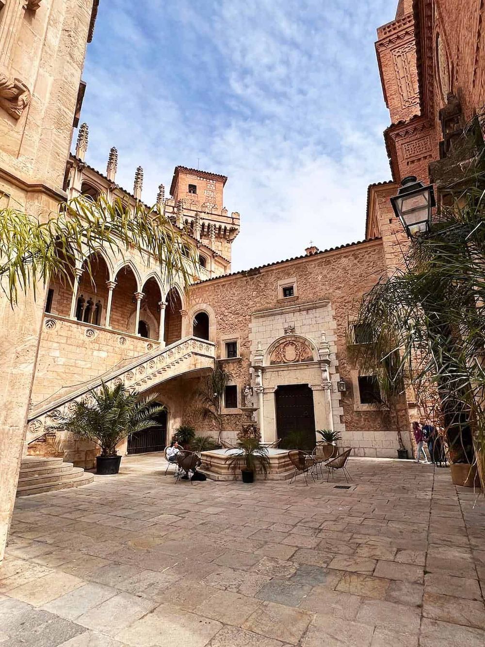 Elegant historic castle courtyard with sandstone architecture, outdoor seating, lush plants, and bright blue sky.