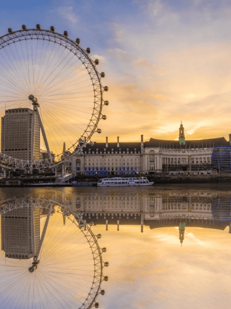 Bright London skyline with London Eye ferris wheel, historic buildings, and sunset reflections over the Thames River.