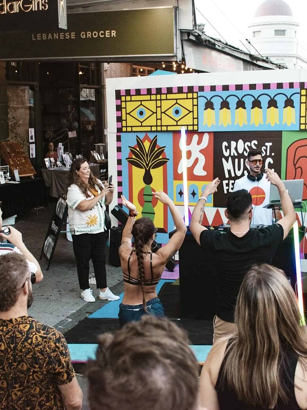 Vibrant street music festival with DJ, lively crowd, outdoor setting, colorful artwork, and audience dancing.