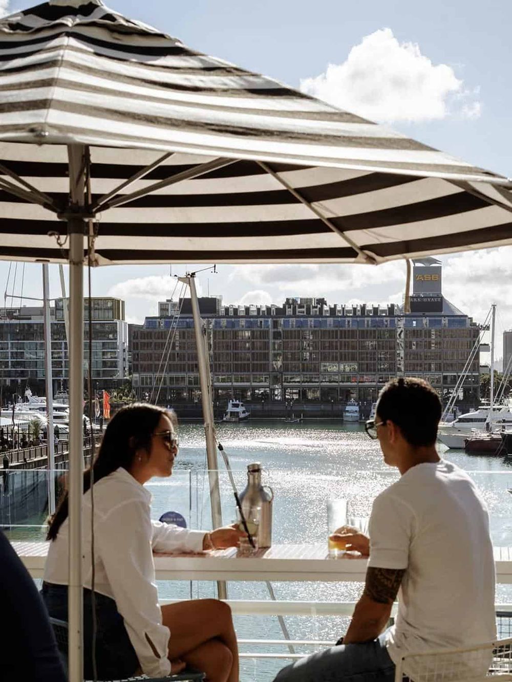 Relaxed couple enjoying drinks under an umbrella near a marina with city buildings in the background, perfect for waterfront dining and navigation.