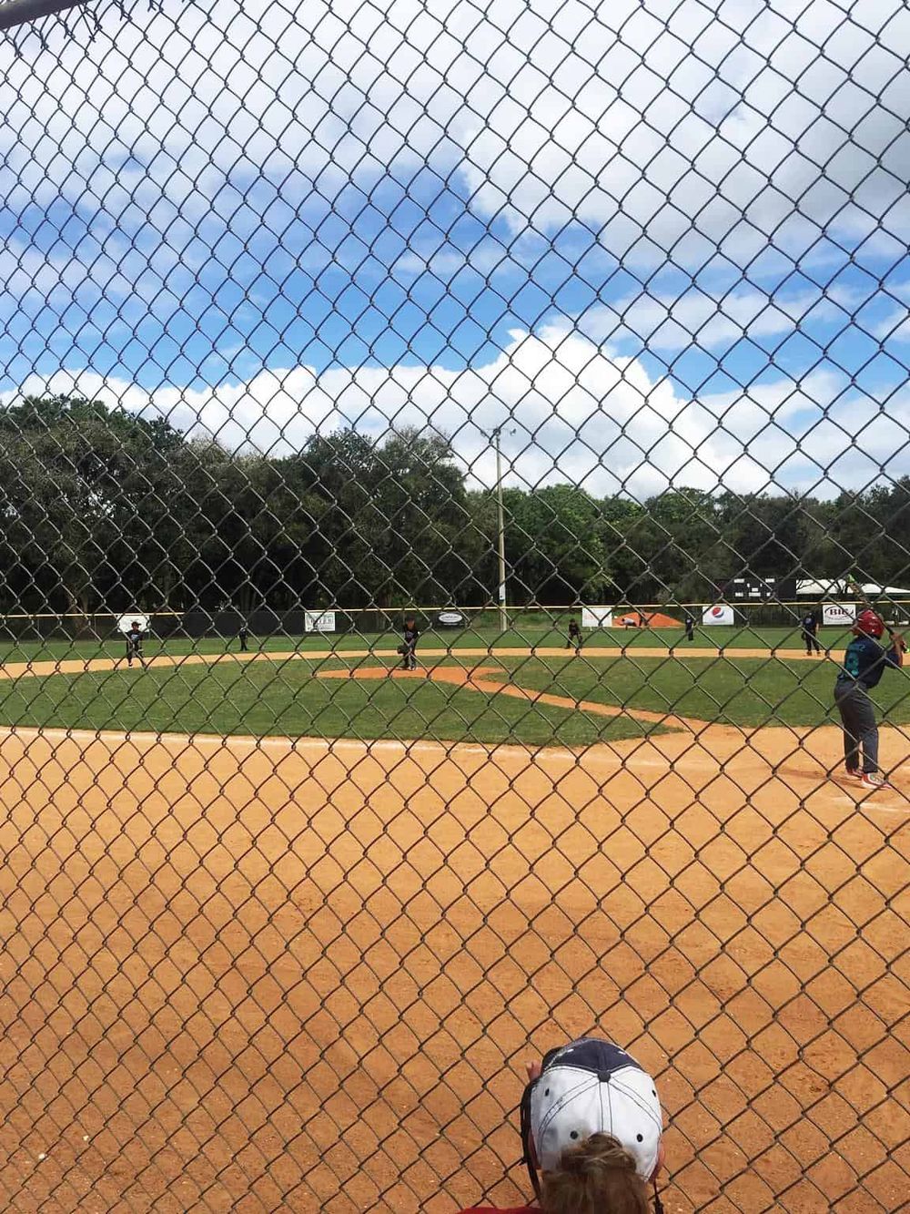 Youth baseball game at local field with chain-link fence in the foreground, players actively engaged on the field.
