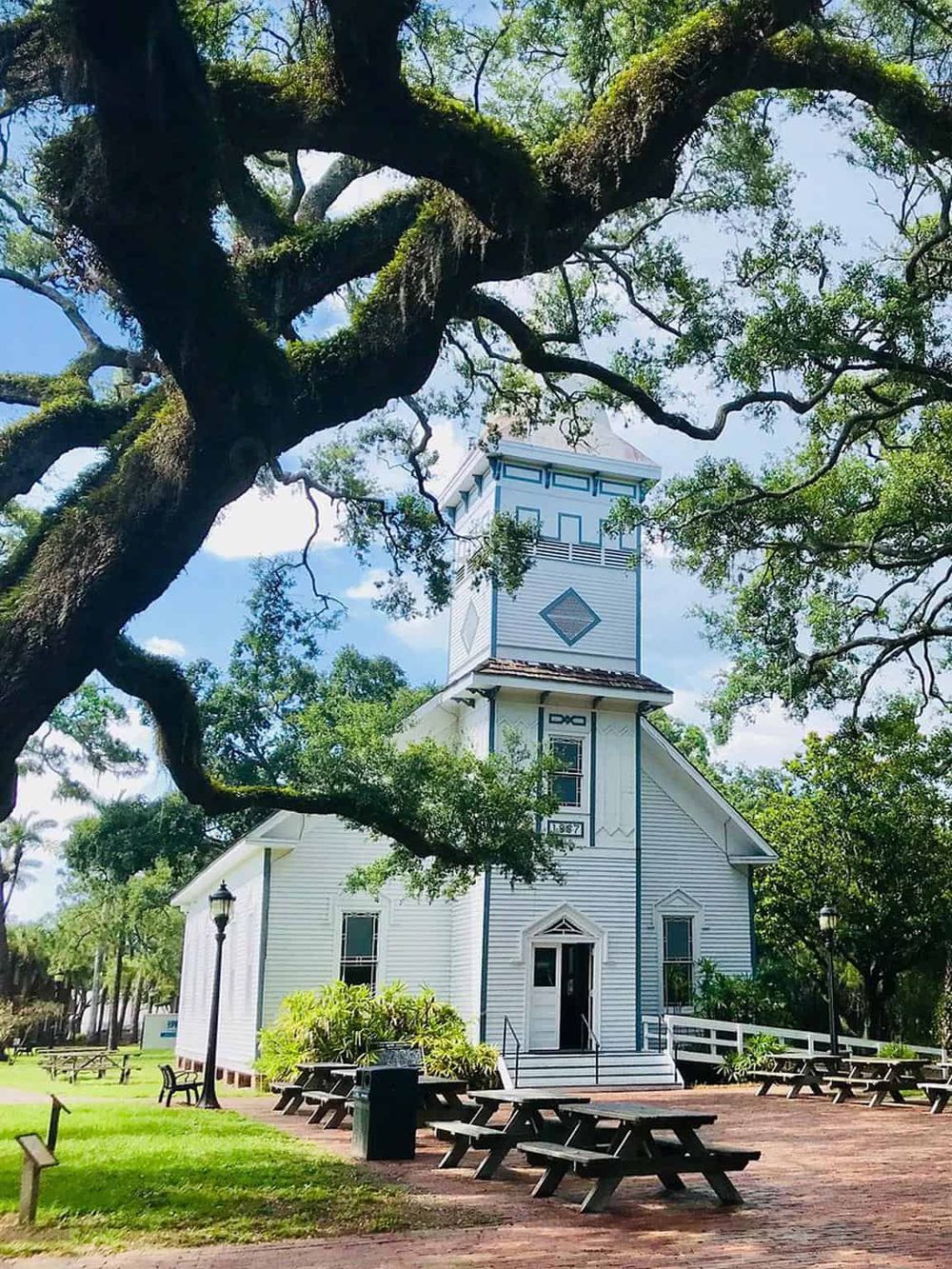 Quaint historic church with a tall steeple surrounded by lush trees and outdoor seating, perfect for community gatherings.