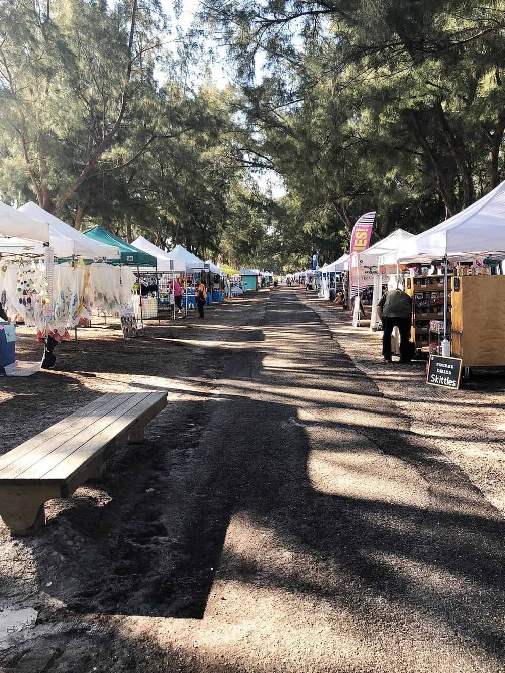 Colorful outdoor market with vendors and tents under tall trees, ideal for exploring local crafts and food.