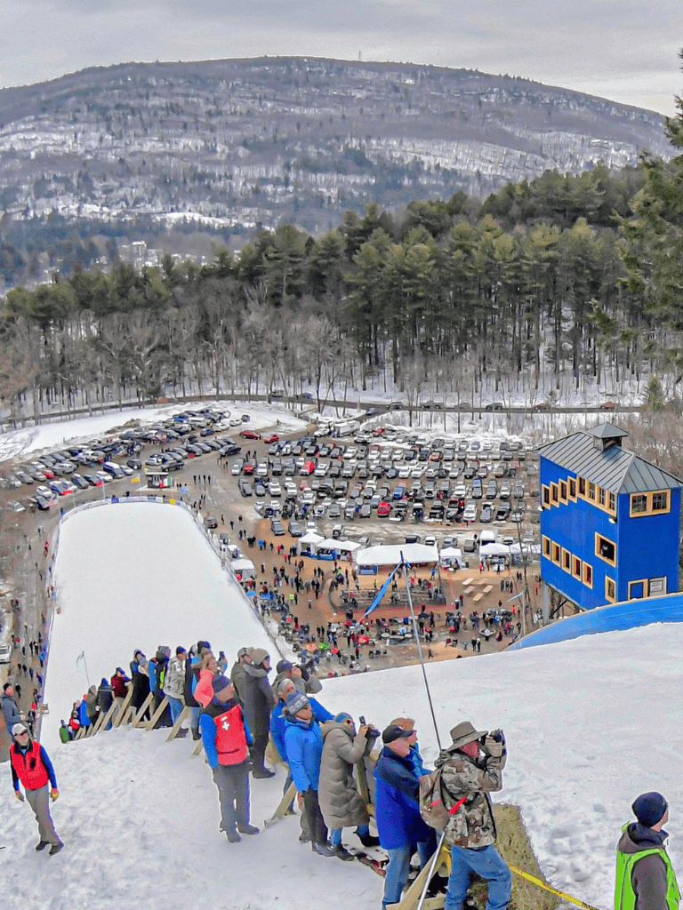 Crowd gathering at the top of a snowy ski jump with parking lot and colorful building in background.