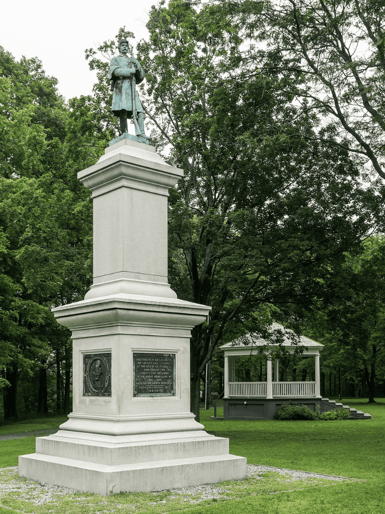 Bronze statue on white monument with greenery background, historic site.