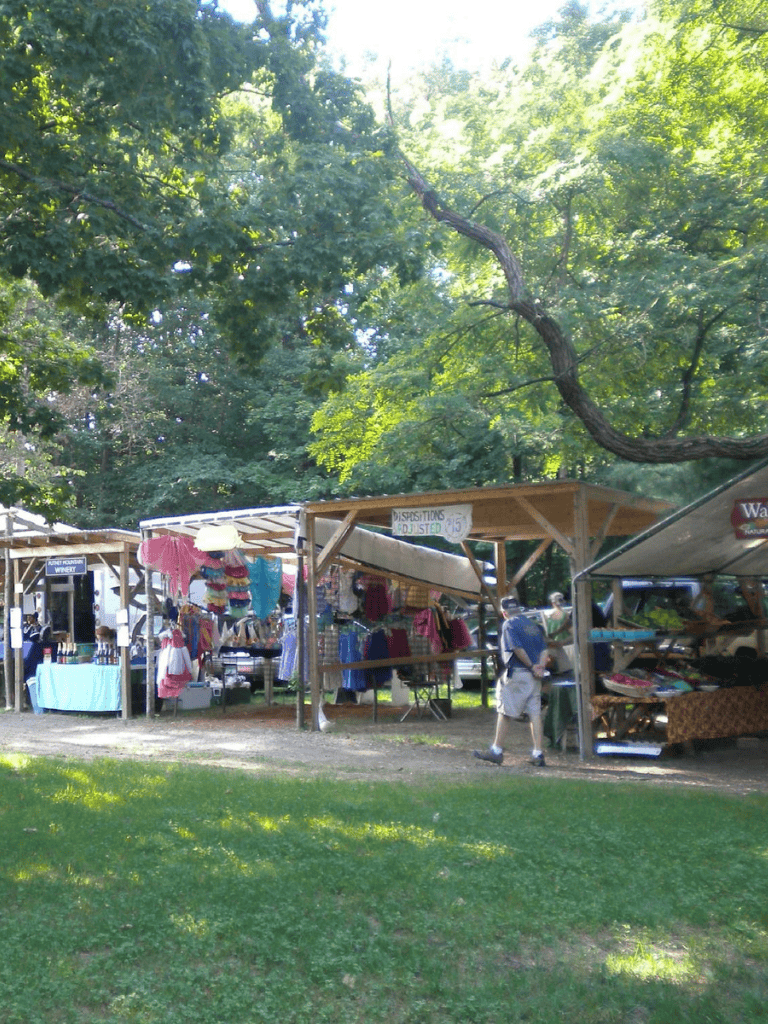 Colorful outdoor craft fair stalls under lush green trees with visitors browsing displays.