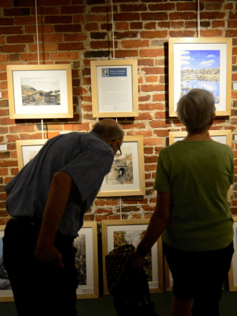 Colorful art exhibition featuring landscape paintings on exposed brick wall with visitors observing art pieces.