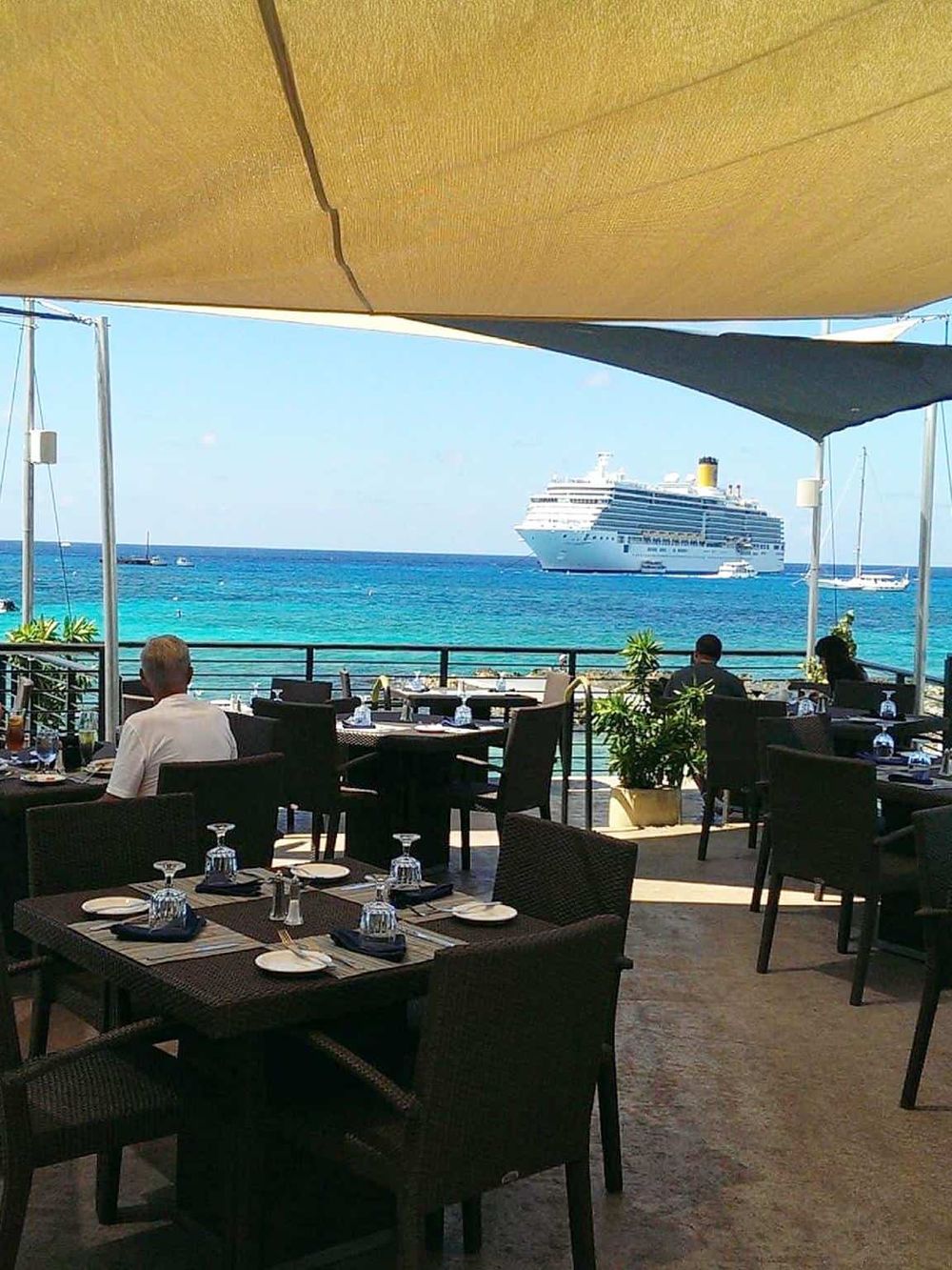 Cruise ship viewed from a seaside restaurant with shaded dining area and ocean scenery.