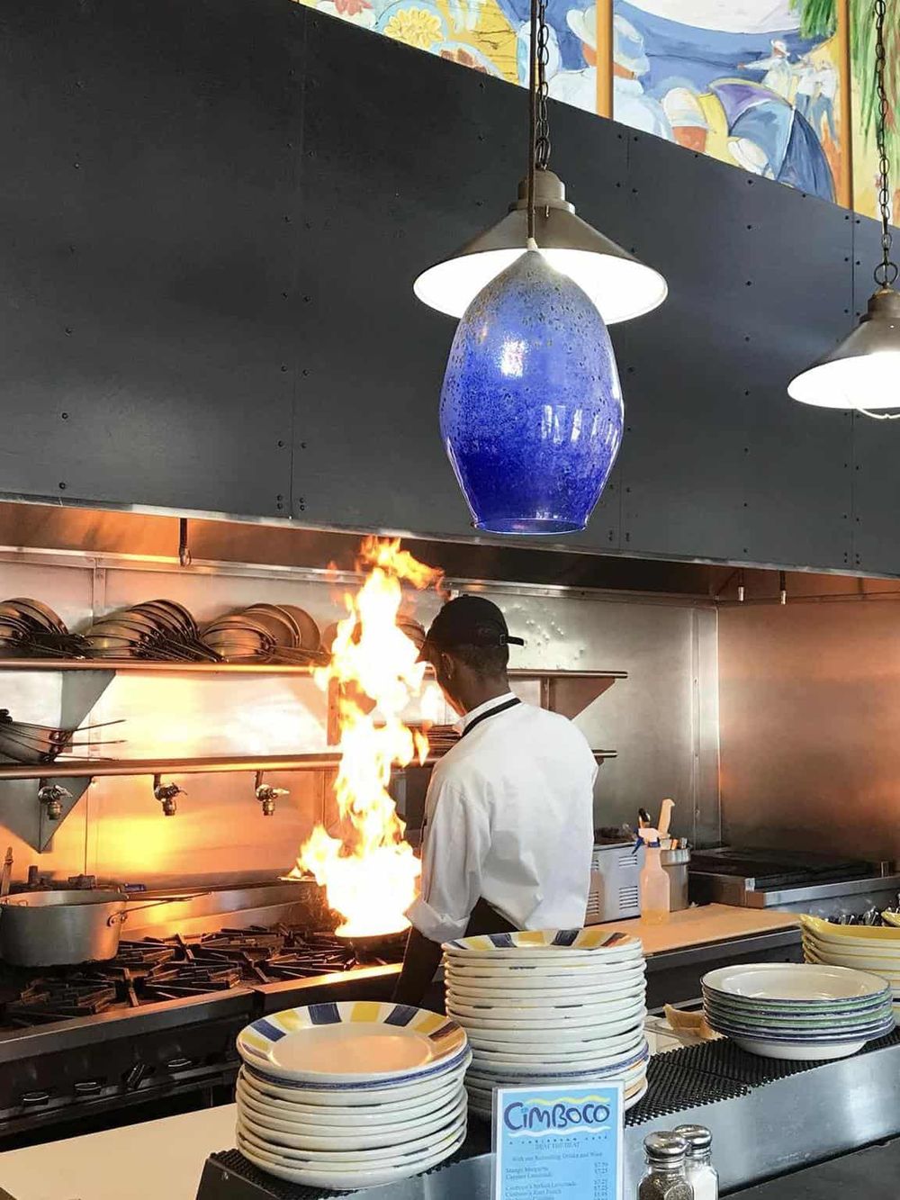 A chef flares flames in a vibrant restaurant kitchen, showcasing professional culinary skills and a lively dining atmosphere.