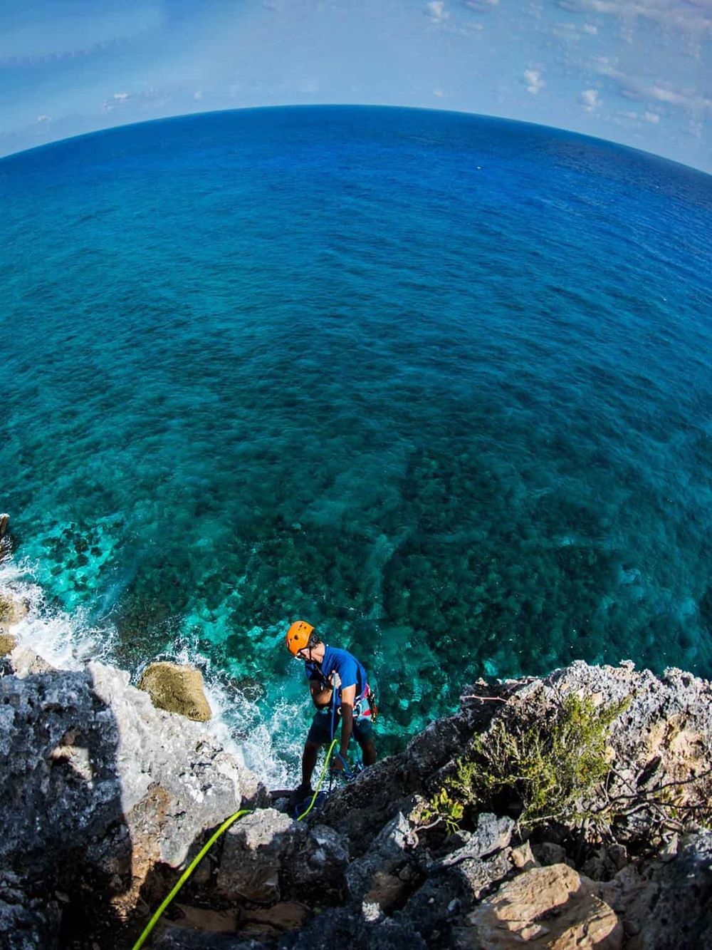 Aerial view of climber rappelling down rugged cliff overlooking bright blue ocean water.