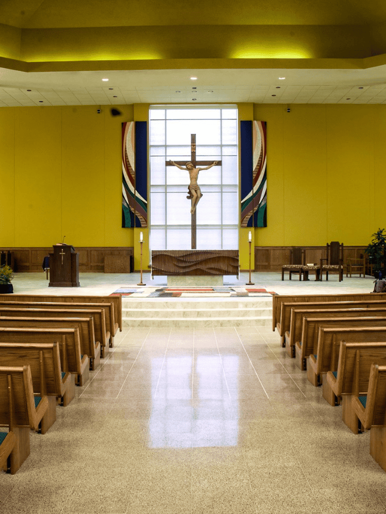 Bright church interior featuring a large crucifix, wooden pews, and vibrant wall art behind the altar.