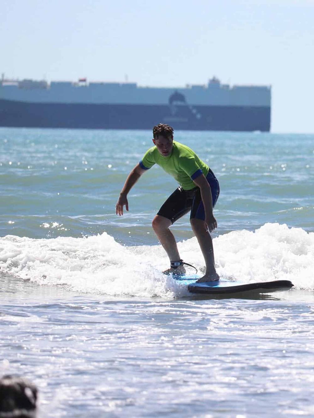 Young man surfing on the beach with a cargo ship in background, adventure travel and ocean exploration.