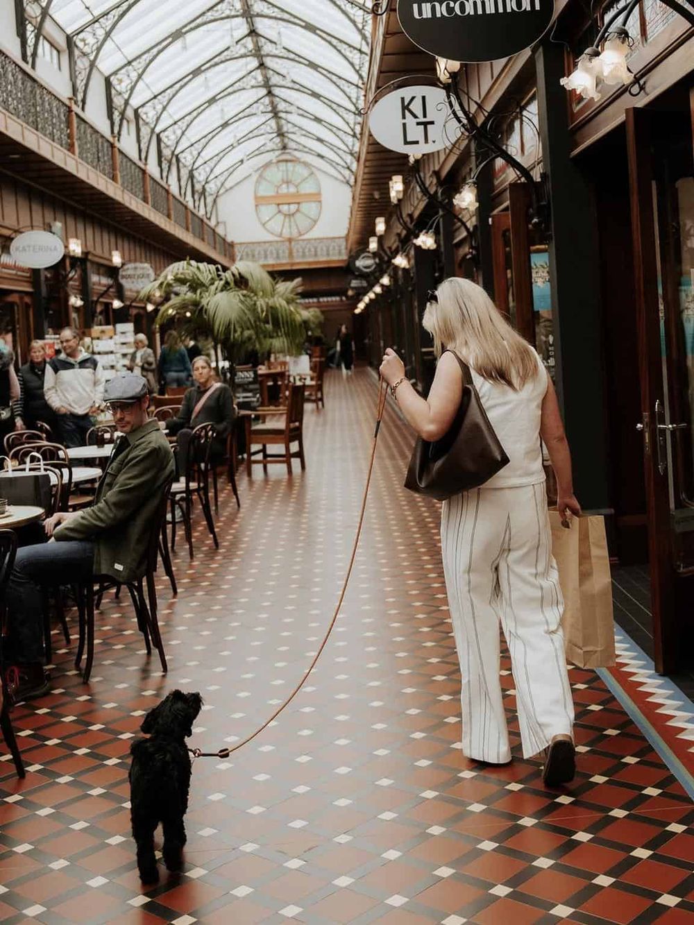 Charming woman walking her small black dog inside an elegant indoor shopping arcade.