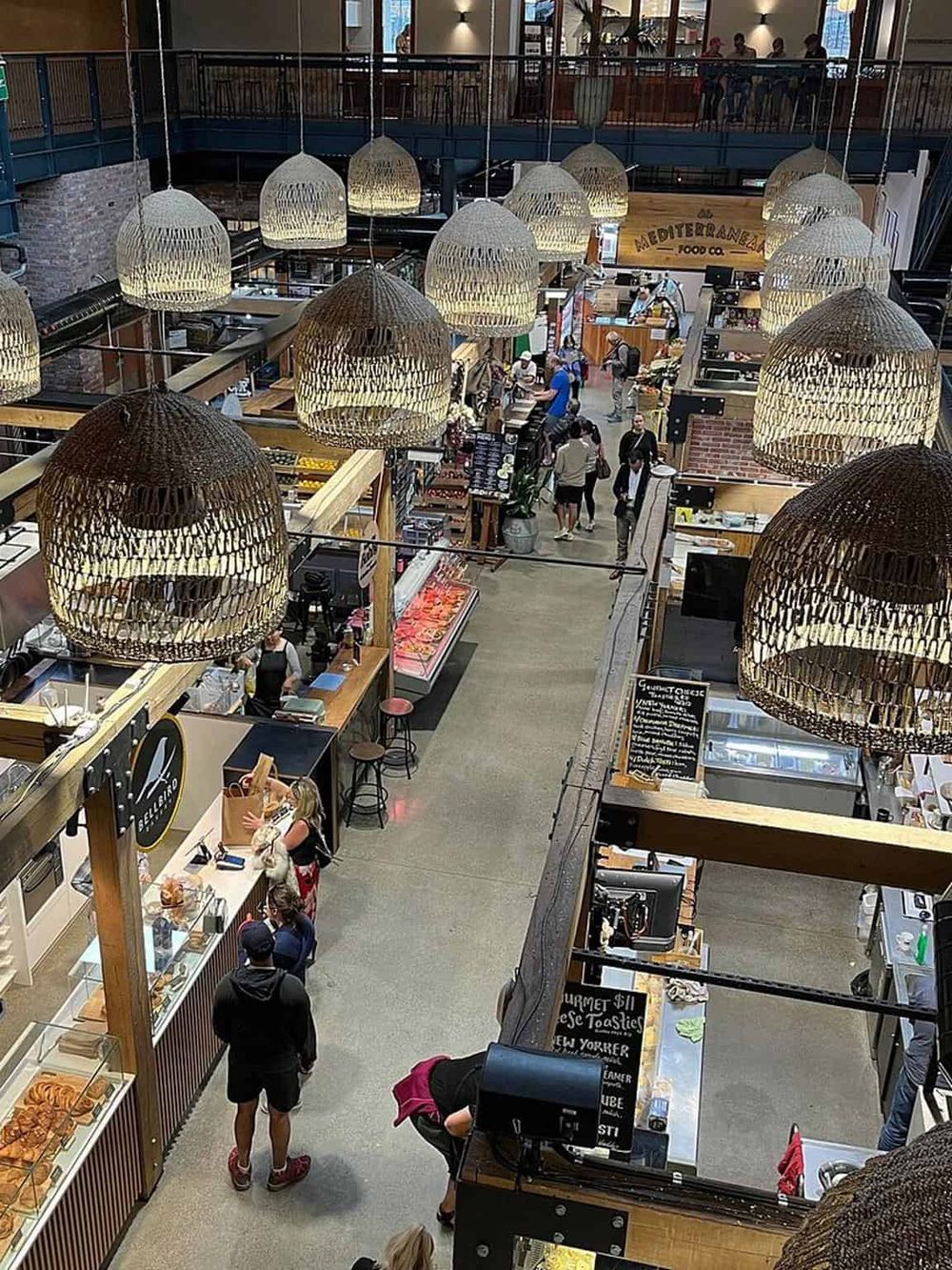 Vibrant indoor marketplace with food stalls, shoppers, and woven pendant lights creating a lively shopping atmosphere.